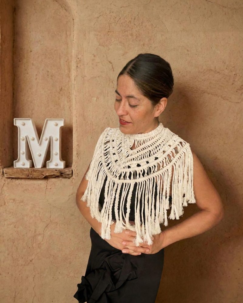 Mujer de cabello oscuro frente a una pared beige, con top de macramé color hueso sobre ropa negra y sus manos unidas cerca de una letra "M" blanca en una repisa de madera.